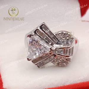 May include: A silver ring with a large triangular-shaped clear gemstone surrounded by smaller clear gemstones. The ring has a unique design with multiple bands and a cluster of smaller stones.