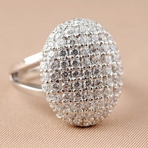 May include: A silver ring with a large, round, oval-shaped center stone. The stone is covered in small, clear, sparkling rhinestones.