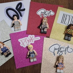 May include: Six plastic minifigures representing musical artists, each mounted on a square glittery card. The cards feature names or logos including Taylor Swift, Beyonce, and Pink. Each minifigure holds a small black microphone accessory.