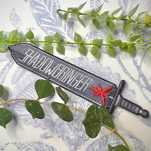 Könnte beinhalten: Ein grauer, gestickter Aufnäher in Schwertform mit dem weißen Text "SHADOWBRINGER". Ein roter Stern ist in der Nähe des Griffs gestickt. Der Aufnäher befindet sich vor einem gemusterten Hintergrund mit grünen Ranken.