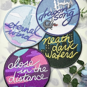 May include: Four embroidered patches with fantasy designs. Each circular patch features a different colour scheme and text: "Eternal Wind," "Dragon Song," "Beneath Dark Waters," and "Close in the Distance." The patches have a stitched border.