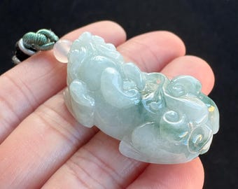 May include: A light green jade pendant carved in the shape of a mythical creature. The jade has areas of darker green and is attached to a green and black cord. The pendant is held in a hand.