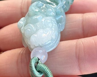 May include: A carved jade pendant in shades of pale green and white, depicting a mythical creature. The pendant is attached to a green cord with a small, round, lavender bead. The cord transitions to a black and white tassel.