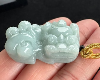 May include: A light green jade pendant carved in the shape of a mythical creature. The pendant has a smooth, polished surface and is attached to a gold-colored cord. The creature has a detailed face and body.