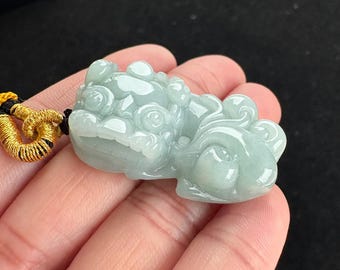 May include: A light green jade pendant carved in the shape of a mythical creature. The pendant has intricate details and is attached to a gold and black cord. The jade has a translucent quality.