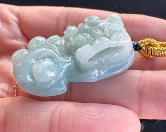 May include: A jade pendant carved in the shape of a mythical creature. The pendant is a pale green color with intricate details and a polished finish. It is suspended from a gold-colored cord.