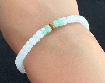 May include: A white beaded bracelet with a mix of light green and brown beads. The bracelet is worn on a wrist, showcasing the delicate design and color contrast. The beads appear to be small and evenly spaced.