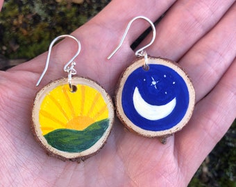 May include: A pair of wooden earrings with hand-painted designs. One earring features a yellow sun rising over a green hill, while the other earring features a blue crescent moon with white stars.