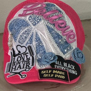 May include: A hot pink trucker hat with a white front panel and a pink mesh back. The hat features the word "Believe" in pink script, denim and sequin accents, and patches that say "Love is in the Hair" and "All Black Everything."