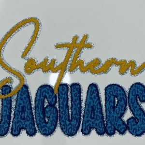 May include: The words "Southern JAGUARS" are displayed in a stylized design. "Southern" is in yellow with silver glitter trim, while "JAGUARS" is in blue with a leopard print pattern and silver glitter trim. The background is white.