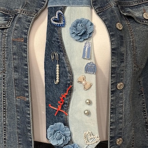 May include: A necktie crafted from light and dark blue denim fabric, featuring a variety of embellishments including fabric flowers, a blue heart pin, a pearl-accented safety pin, a red script word faith, and several small metal pins.