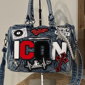 May include: A blue acid-wash denim handbag featuring the word ICON in large red, black, and white letters. The bag is decorated with various patches including a turntable, a DJ, a red cross, and the word Vintage, plus a white pearl chain strap.