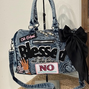 May include: A blue acid-wash denim handbag featuring a large black patch that reads "blessed all the time," a "NO is a full sentence" patch, an "Old School" patch, a "Clock That Tea" patch with a hand graphic, and a large black satin bow.