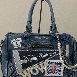 May include: Denim handbag with Seattle Seahawks team logo, embellished with rhinestones and pearls. Features a "WOW" pearl accent and "Gameday" patch. Includes a blue flower and a chain strap.