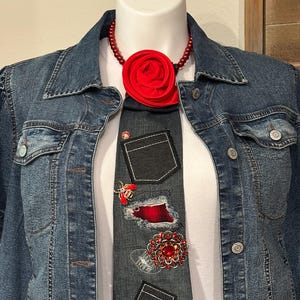 May include: A dark gray denim necktie featuring two small patch pockets, a red fabric rose at the collar, a red bee pin, a red plaid fabric patch with frayed edges, and a large circular brooch with red crystals, worn over a white shirt and denim jacket.