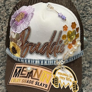 May include: A brown trucker hat with a white front panel, embellished with embroidered flowers, rhinestones, and the word "Baddie". The hat features the text "MEAN IN EVERY SHADE SLAYS" and "music is my life".