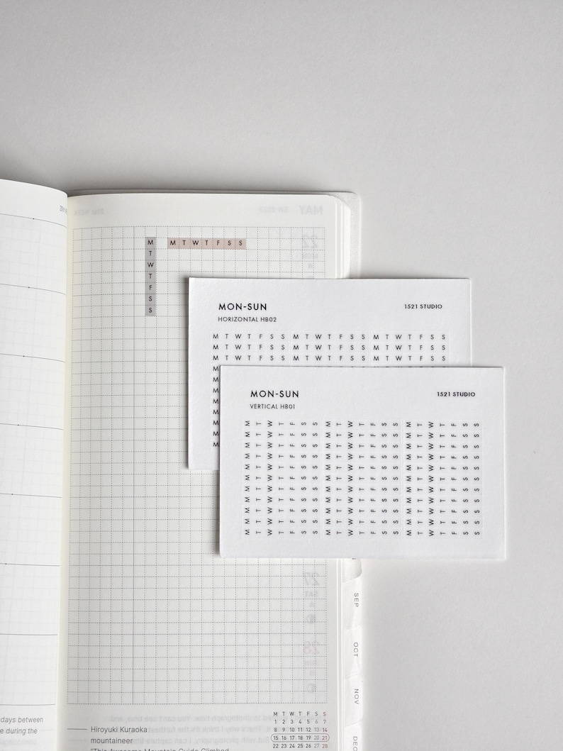 May include: An open planner with a grid layout featuring two white rectangular stickers. The top sticker reads MON-SUN HORIZONTAL HB02 with rows of M T W T F S S. The bottom sticker reads MON-SUN VERTICAL HB01 with columns of M T W T F S S.