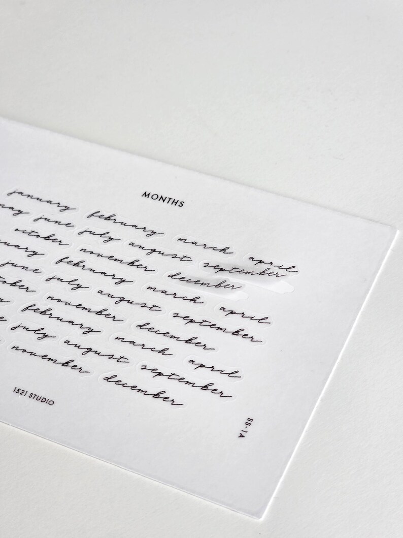 May include: A clear rectangular sticker sheet featuring the word MONTHS in a bold sans-serif font at the top, followed by multiple rows of the twelve months of the year written in a black cursive script. The bottom corners display 1521 STUDIO and SS-1A.