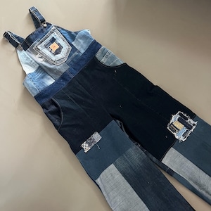 May include: Denim overalls featuring a patchwork design with varying shades of blue and black fabric. The garment includes a chest pocket with a small orange label, adjustable shoulder straps, side pockets, and decorative patches on the legs.