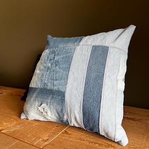 Pode incluir: Uma almofada quadrada de ganga com um design de patchwork. A almofada apresenta várias tonalidades de ganga azul, incluindo lavagens claras e escuras, dispostas num padrão de riscas e painéis. A almofada está colocada numa superfície de madeira.