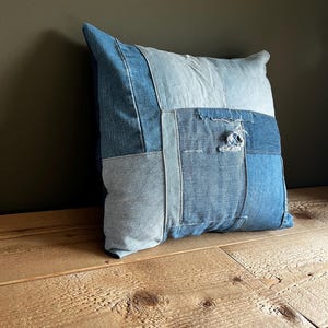 Può includere: Cuscino quadrato in denim con un design patchwork. Il cuscino presenta varie tonalità di denim blu, creando una superficie strutturata e visivamente interessante. Il cuscino è appoggiato su una superficie di legno.