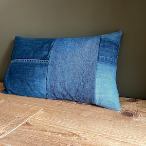 Może przedstawiać: Prostokątna poduszka z denimu z patchworkowym wzorem. Poduszka wykonana jest z różnych odcieni niebieskiego denimu, z widocznymi detalami szwów. Leży na drewnianej powierzchni na tle ciemnozielonego tła.