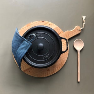 May include: A black cast iron Staub Dutch oven with a denim pot holder sits on a round wooden chopping board. A wooden spoon is to the right. The Dutch oven has the word "STAUB" on the side.