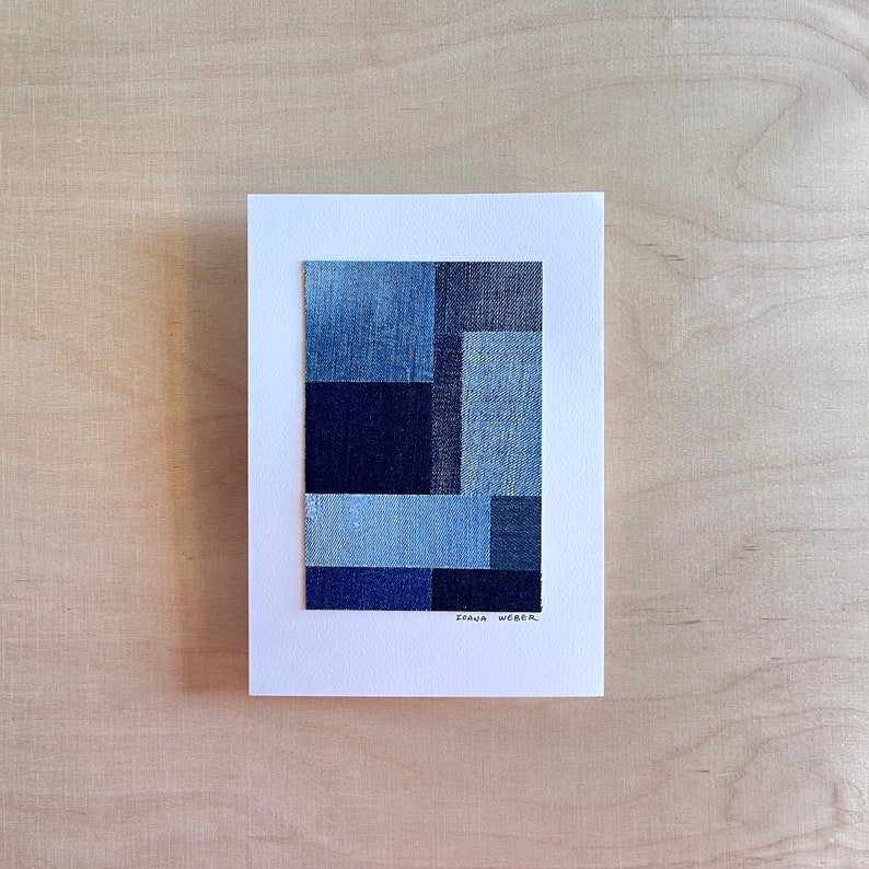 May include: A framed abstract art piece made of denim fabric in shades of blue. The artist's signature, "ISANA WEBER", is written in black ink at the bottom of the piece.