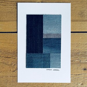 May include: A framed abstract art print featuring a geometric design made of different shades of blue denim fabric. The print is signed "Ioana Weber" in the bottom right corner.