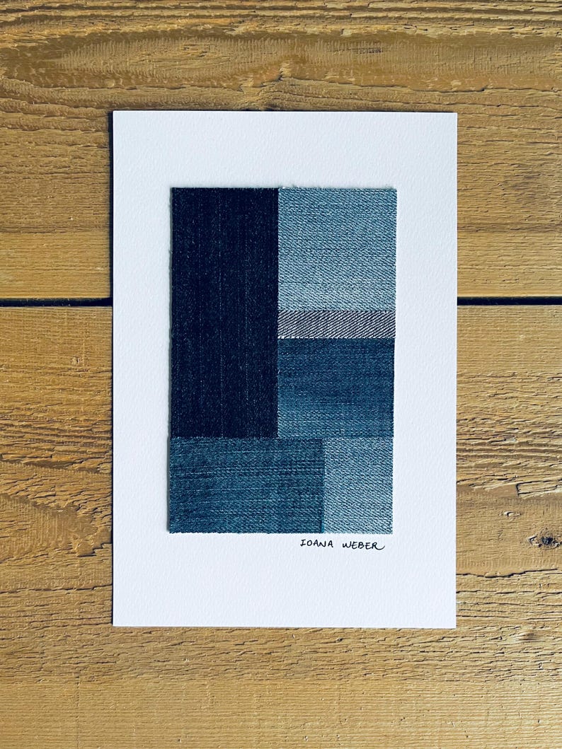 May include: A framed abstract art print featuring a geometric design made of different shades of blue denim fabric. The print is signed "Ioana Weber" in the bottom right corner.
