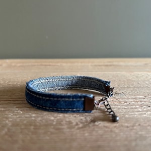 Puede incluir: Un brazalete de denim azul con un cierre y una cadena plateados. El brazalete está sobre una superficie de madera.