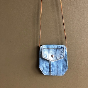 May include: A small blue denim crossbody bag with a brown leather strap. The bag has a flap with a single button closure and a small pocket on the front.