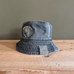 Könnte beinhalten: Ein schwarzer Bucket-Hut aus Denim mit einem runden Patch, der auf die Krone genäht ist. Der Patch besteht aus einem anderen Material und hat eine gelbe Naht um den Rand.