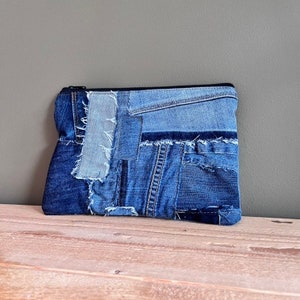 Puede incluir: Un bolso de mano rectangular de mezclilla con un diseño de patchwork. El bolso está hecho de varios tonos de mezclilla azul, con costuras visibles y un cierre de cremallera negro. La bolsa está sobre una superficie de madera.