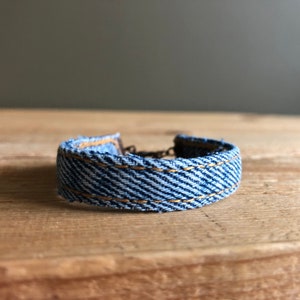 Puede incluir: Un brazalete de mezclilla azul con costuras doradas. El brazalete está hecho de mezclilla reciclada y es una excelente manera de agregar un toque de estilo a cualquier atuendo.