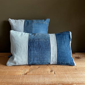 Puede incluir: Dos cojines rectangulares de mezclilla con un diseño de patchwork. Los cojines presentan varios tonos de mezclilla azul, con secciones azul claro y azul medio. Se colocan sobre una superficie de madera contra una pared marrón oscuro.
