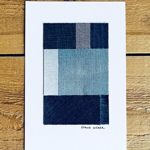 May include: A framed abstract art print featuring a geometric design of four squares made from different shades of blue denim fabric. The artist's signature, "Ioana Weber", is written in black ink at the bottom of the print.