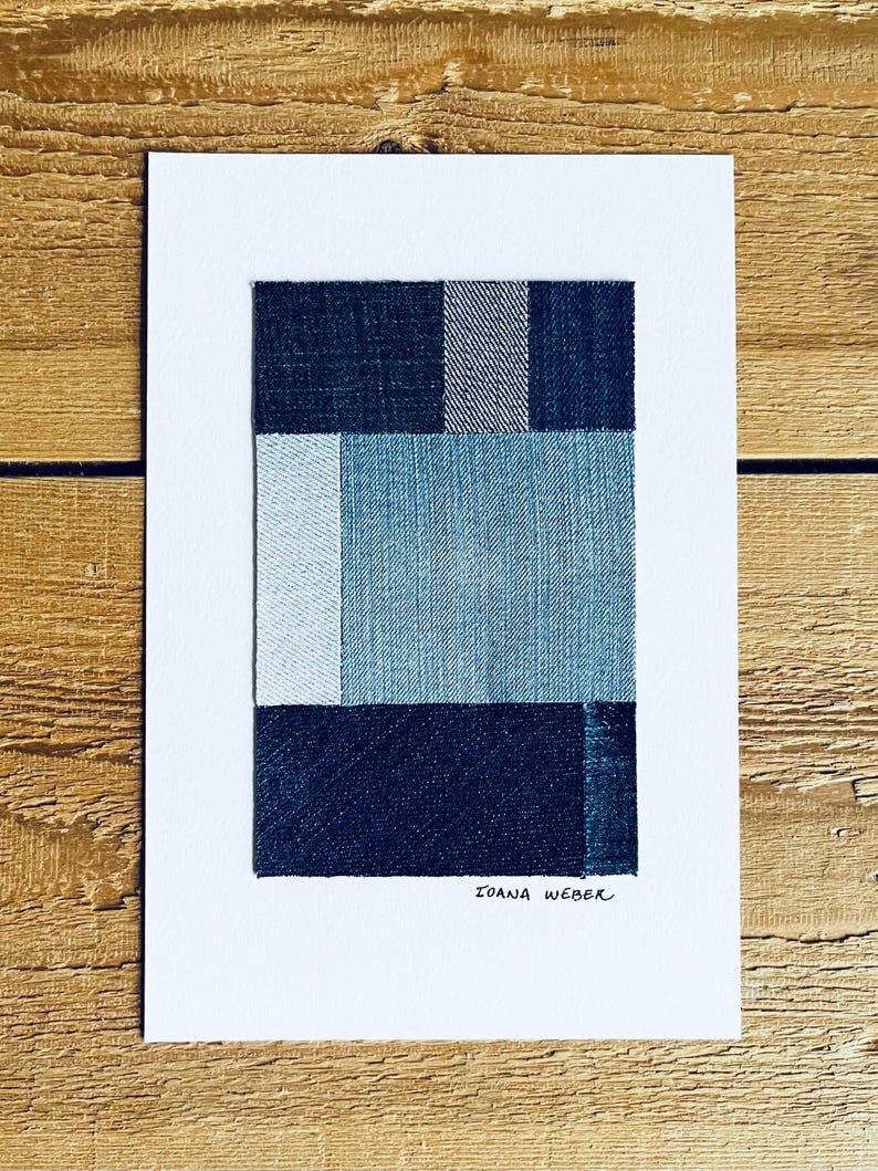 May include: A framed abstract art print featuring a geometric design of four squares made from different shades of blue denim fabric. The artist's signature, "Ioana Weber", is written in black ink at the bottom of the print.
