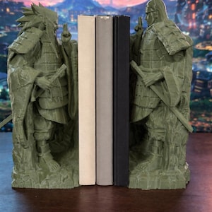 May include: Green statue bookends depicting two figures in armour, flanking three books. The bookends have a detailed design, and the books are beige, grey, and black. The background shows a fantasy landscape.