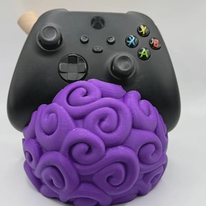 May include: A black Xbox controller rests on a vibrant purple, swirled, spherical object. The controller has colored buttons and directional controls. The purple object has a unique, textured design, resembling a stylized fruit or orb.