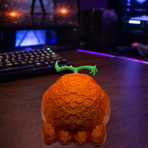 May include: An orange, textured fruit with a green stem, resembling a devil fruit from the anime series. The fruit is on a wooden surface, with a computer and keyboard in the background.