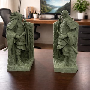 May include: Two sage green samurai warrior bookends. Each bookend depicts a detailed figure in traditional armour, holding a sword. The figures stand on a textured base, adding depth. The bookends are placed on a dark wooden surface.