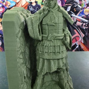 May include: A green 3D-printed figurine of a warrior in armour, standing on a rocky base. The figure is detailed with segmented armour, a helmet, and a sword. The background features colourful anime-style characters.