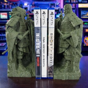 May include: Green samurai bookends supporting three PS5 game cases. The games are "The Last of Us Part II Remastered", "Final Fantasy VII Rebirth", and "Ghost of Yotei". The bookends feature detailed samurai armour.