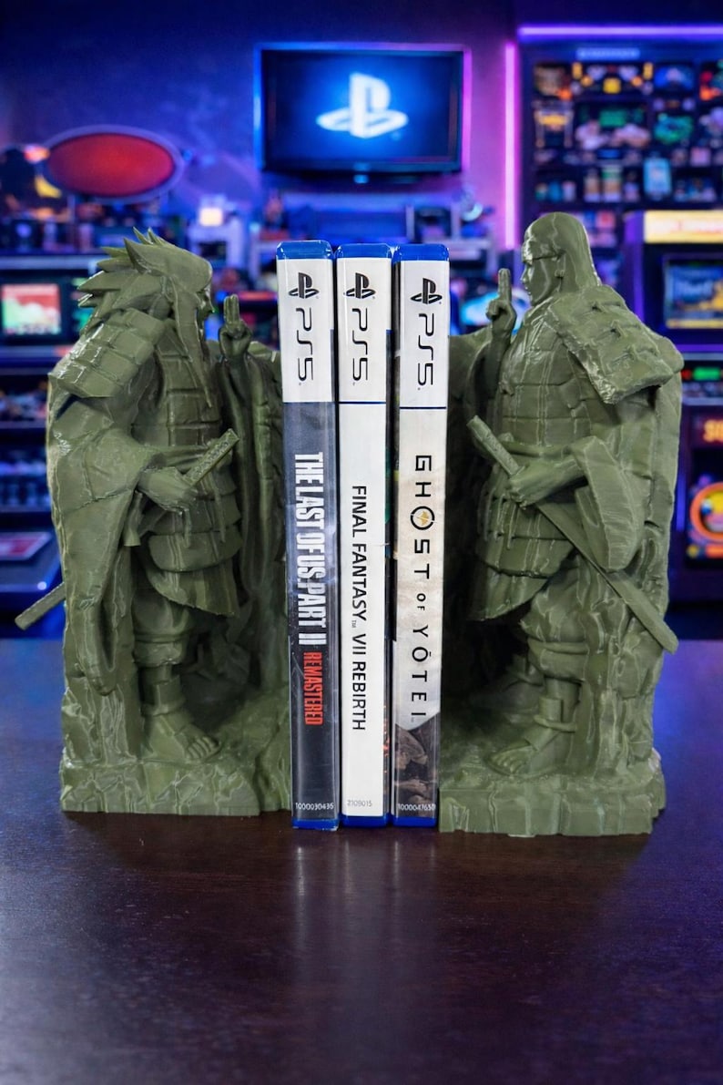 May include: Green samurai bookends supporting three PS5 game cases. The games are "The Last of Us Part II Remastered", "Final Fantasy VII Rebirth", and "Ghost of Yotei". The bookends feature detailed samurai armour.