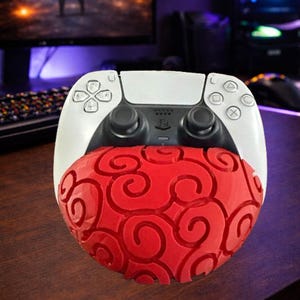 May include: A white and black PlayStation 5 controller with a red, patterned accessory. The controller is on a wooden surface, with a computer monitor and keyboard in the background. The accessory has a swirling design.