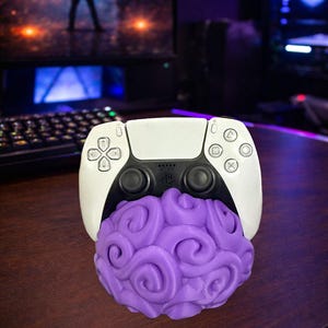 May include: A white and black PlayStation 5 controller with a purple, brain-shaped object attached. The controller has the standard button layout. The background includes computer monitors and a keyboard.