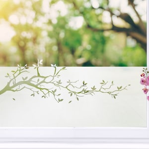 Może przedstawiać: Białe okno z matowym wzorem przedstawiającym zieloną gałąź drzewa z liśćmi. Wazon z różowymi orchideami stoi na parapecie. Tło przedstawia rozmyty zielony krajobraz.
