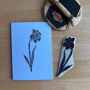 Op de afbeelding: Een zwart-witte bloemenstempel met bijpassende print op een lichtblauwe kaart. Een ronde, geweven handgreep met een zwart oppervlak en Japanse tekst staat op de achtergrond. De stempel en print tonen een narcisontwerp.
