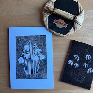 Op de afbeelding: Een stilleven met een lichtblauwe kaart met een zwart-witte bloemenprint, een kleine zwarte kaart met een vergelijkbaar bloemmotief en een rond, geweven object met Japanse tekst.